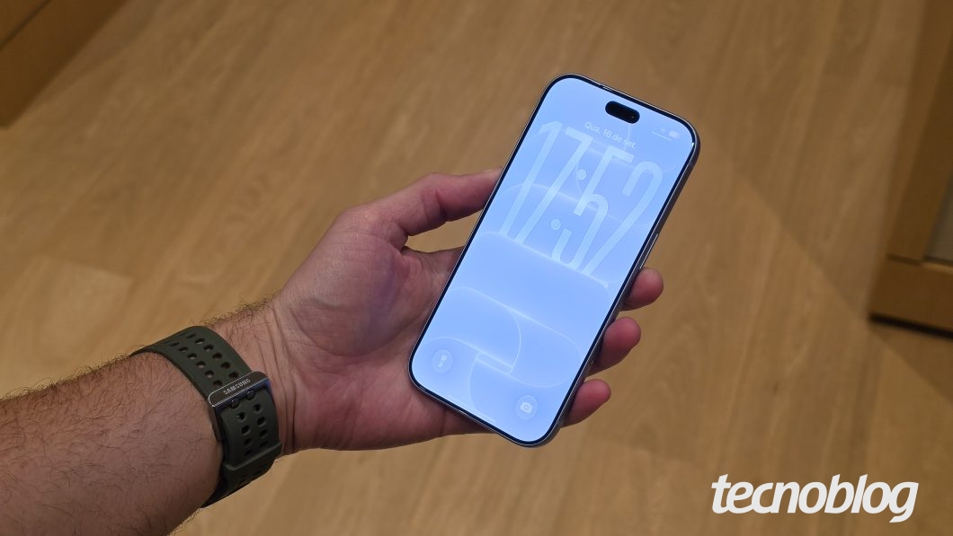 Imagem mostra um iPhone sendo segurado em uma mão. A tela está virada para a câmera, exibindo um relógio com o horário 17:52. Na parte inferior direita, a marca d'água do "tecnoblog" é visível.