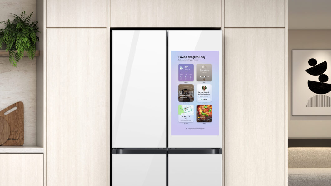 Foto de um refrigerador branco de quatro portas, modelo Samsung Family Hub, embutido em uma cozinha moderna com armários de madeira. A grande tela vertical na porta direita está ligada, exibindo a interface One UI com vários widgets, incluindo clima, calendário e fotos.