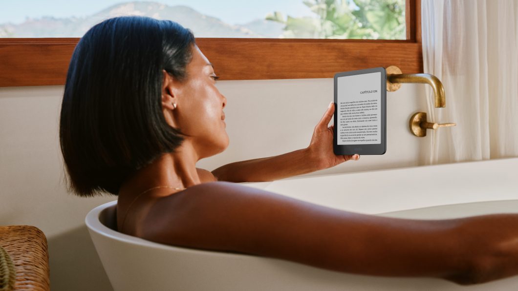 Fotografia de estilo de vida mostra uma mulher de cabelos pretos e curtos vista em perfil dentro de uma banheira branca cheia de água; ela segura com a mão direita um leitor digital Amazon Kindle preto.