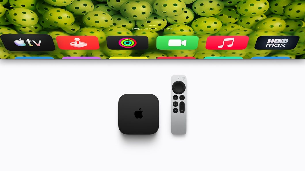 Imagem mostra um Apple TV 4K preto, em formato quadrangular, ao lado de um controle remoto prateado. À frente, uma televisão exibe aplicativos como o Apple Música e o HBO Max