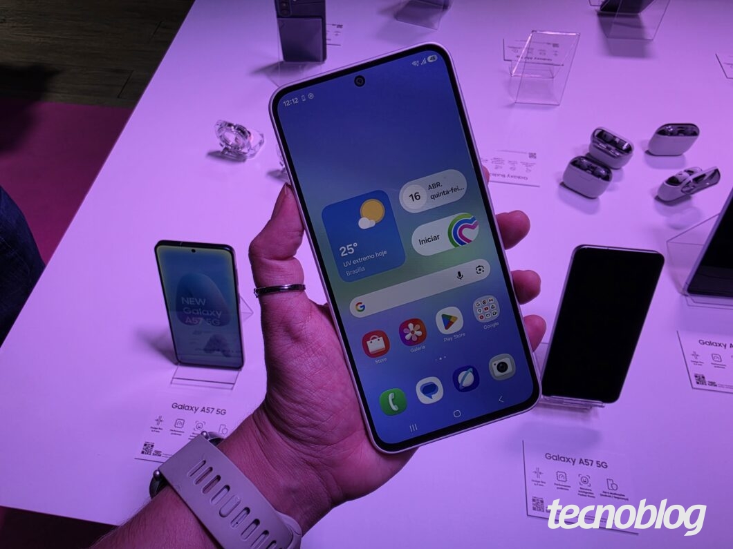Pessoa segurando um smartphone Galaxy com tela inicial aberta, ao lado de embalagens de Galaxy A57 5G