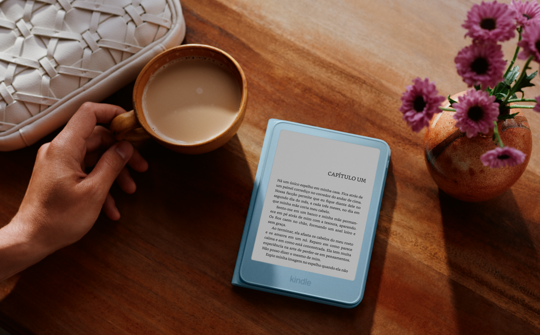 Fotografia mostra uma mesa de madeira rustica, exibindo no centro um leitor digital Amazon Kindle de cor azul-claro com a tela ligada no texto "CAPÍTULO UM"; à esquerda, a mão de uma pessoa segura a alça de uma xícara de cerâmica com café com leite.